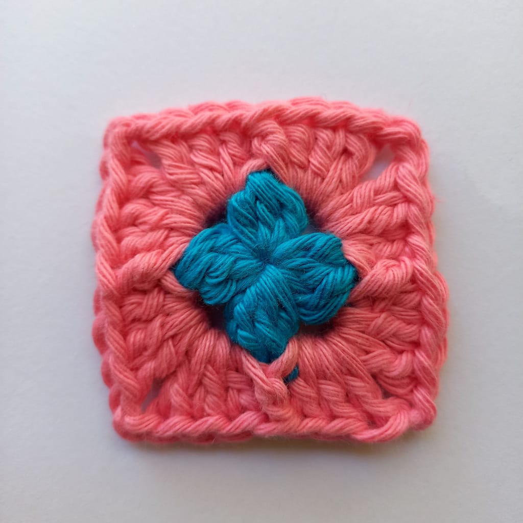 a tiny crochet granny square, the first round is blue yarn and the second round is pink yarn