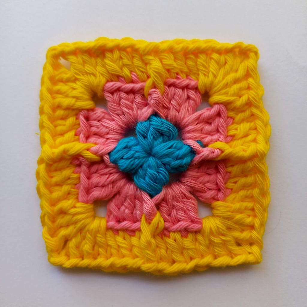 a small textured crochet granny square with a variety of stitches. The first round is blue yarn, the second round is pink yarn and the final round is yellow yarn