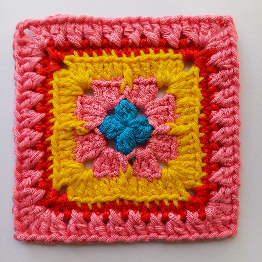 a crochet granny square in pink, blue, yellow and red yarn comprising different types of twisted and raised crochet stitches including crossed double crochet, front posts and back loop only
