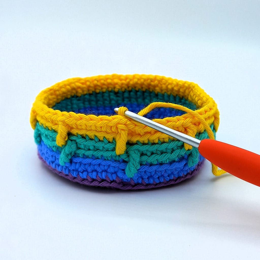 striped small crochet basket in progress, blue, gree and yellow stripes worked by orange crochet hook