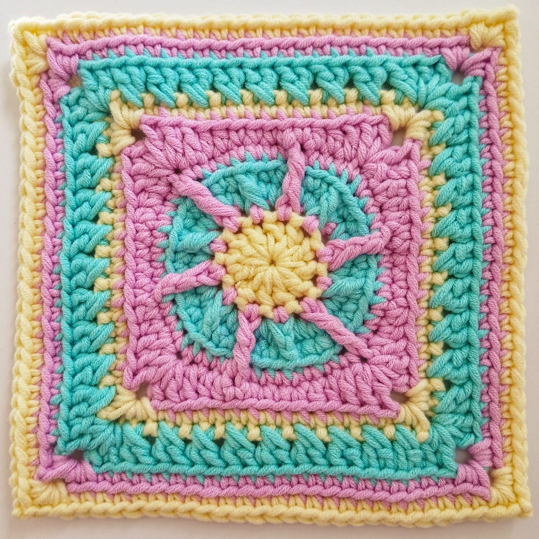 A small, textured pink, yellow and blue granny square here shown at round 10 of the accompanying written crochet pattern instructions