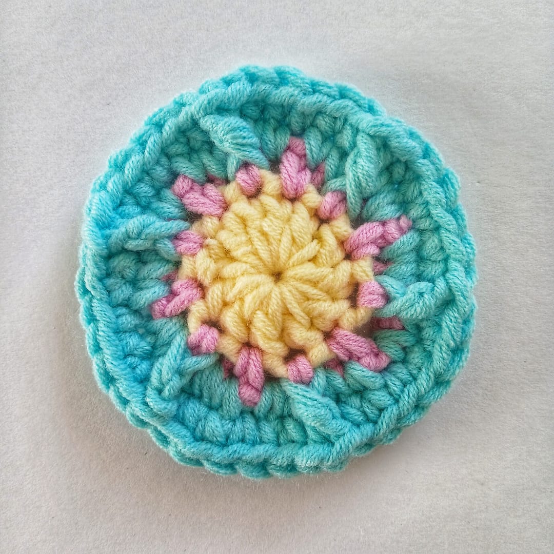 A small crochet circle showing the first 4 rounds of the crochet pattern in yellow, link and blue yarn