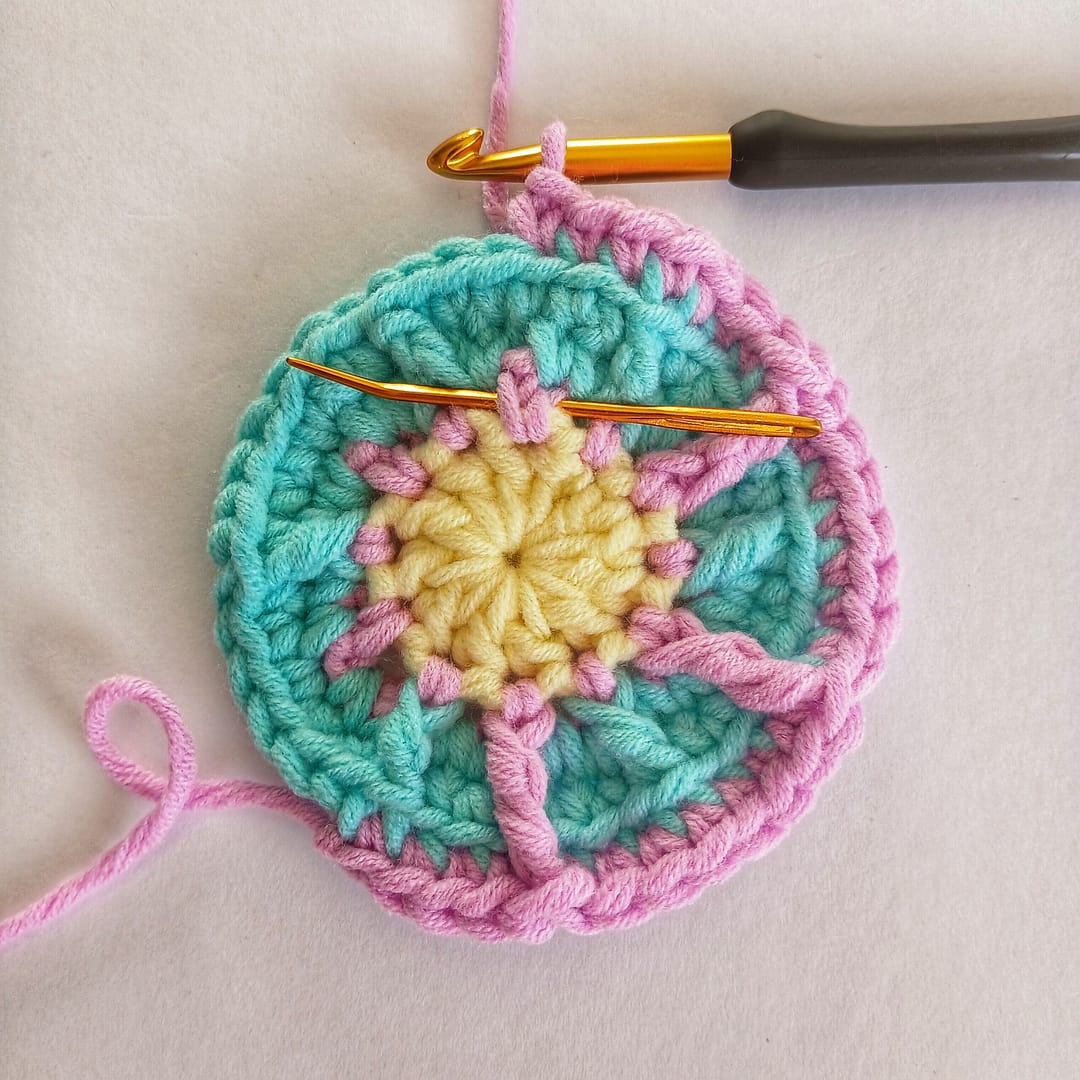 A small crochet circle showing the progress in pink yarn of the fifth round of this pattern with a needle showing the correct placement of the FPtr stitches