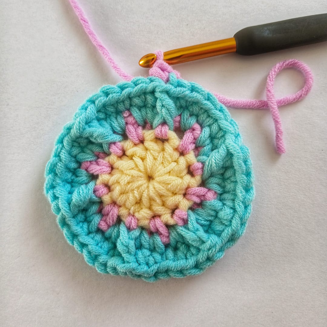 A small crochet circle in yellow, pink and blue yarn with a crochet hook insterted one stitch into the fifth round of this pattern
