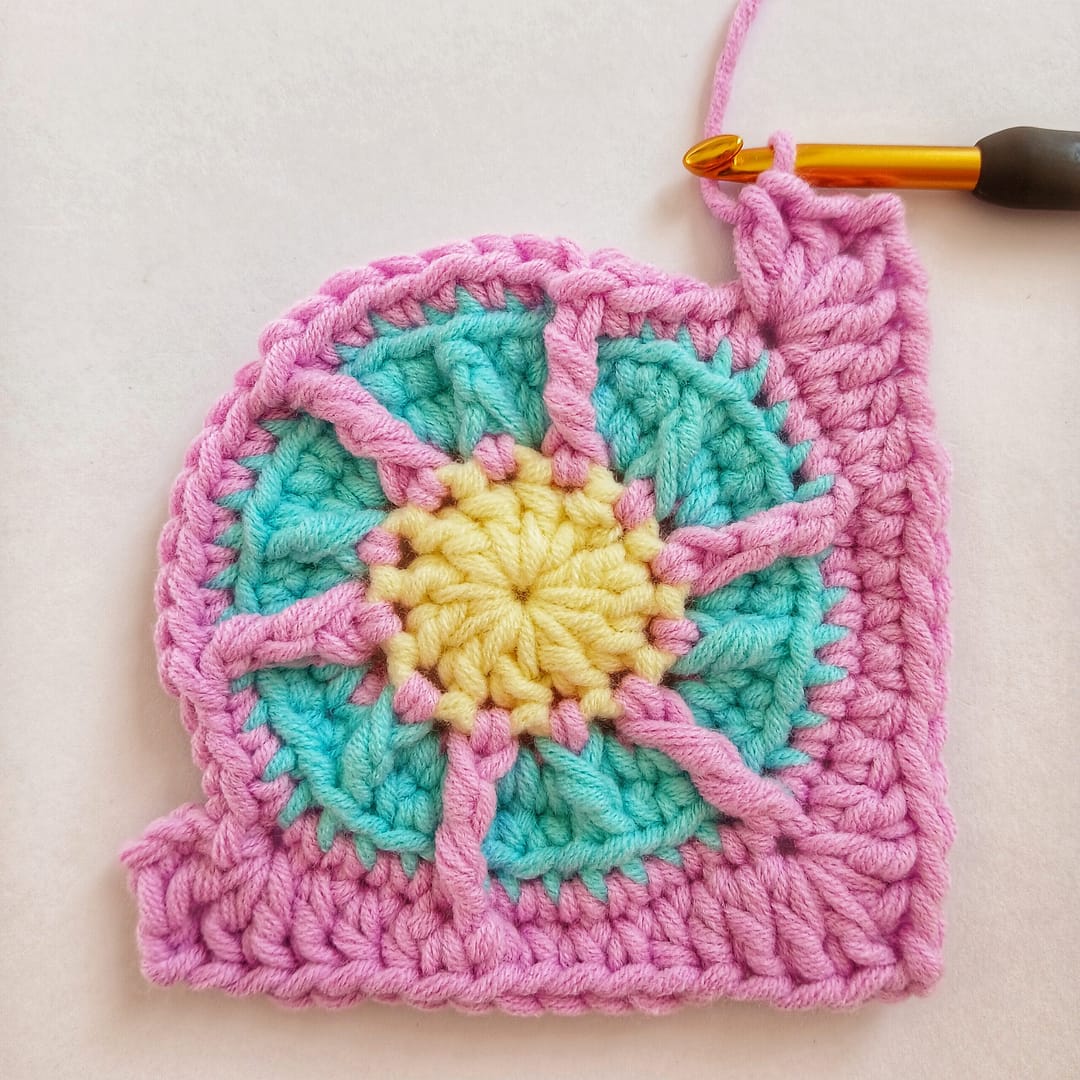 A small crochet circle that is being made into a square in this round of the pattern instructions, picture shows 4 corners being added in pink yarn with a black working crochet hook attached