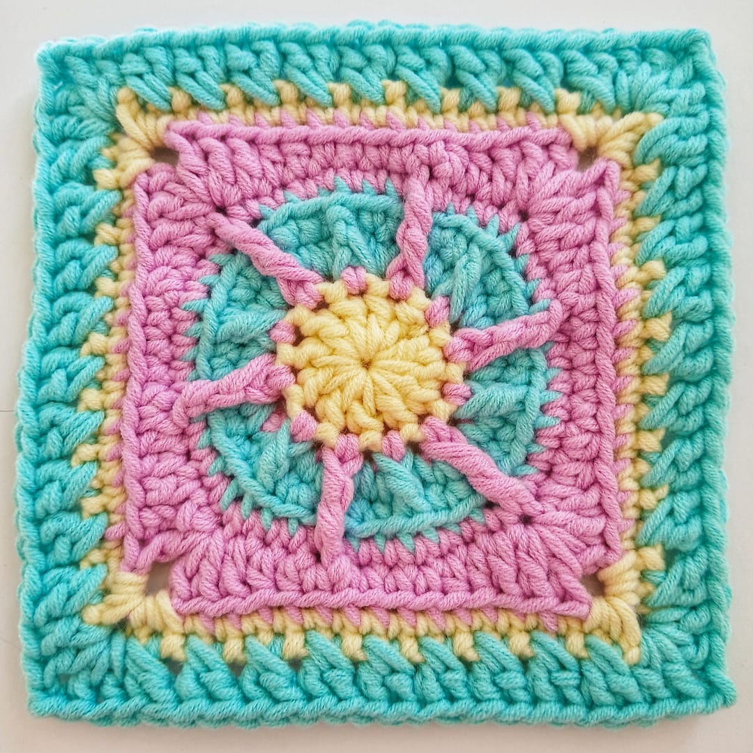 A close up of the granny square as complete at round 8 of the written instructions; colours are pink, yellow and blue in this textured crochet design