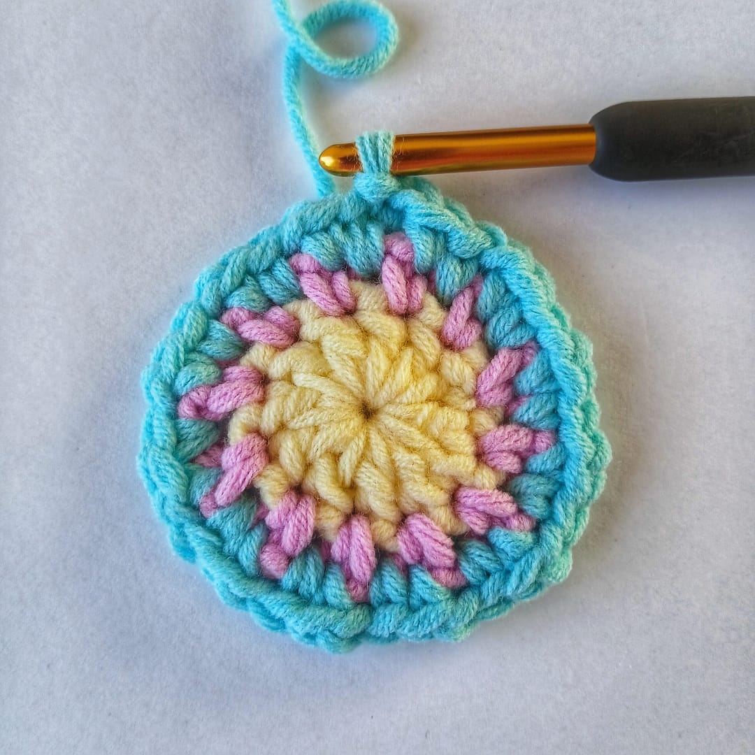 A small crochet circle showing the first 3 rounds of the pattern instructions in this post, using yellow, pink and blue yarn and with a black crochet hook in the final stitch