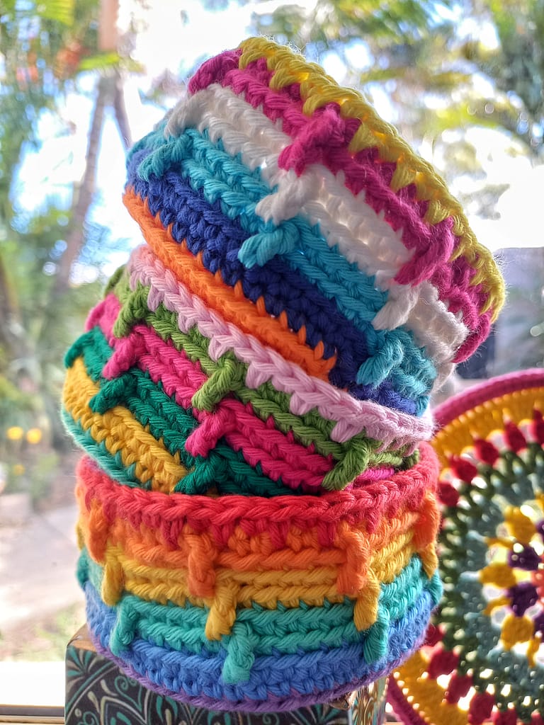 3 stacked small colourful crochet baskets with a mandala half showing in the background and out of focus trees